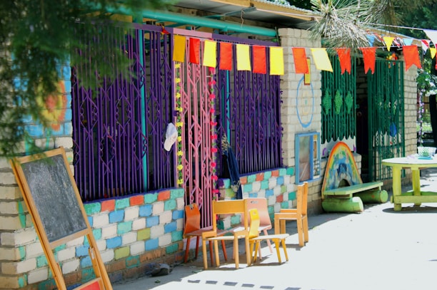 A colorful outdoor setting features a wall painted with alternating blue, white, and pink bricks. Bright metal bars in purple and green cover the windows. A series of colorful triangular flags hang overhead. A small chalkboard and several wooden chairs are arranged near the wall. On the ground, a painted rainbow decorates the side of a small structure next to a circular green table.