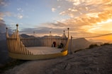 A scenic viewing platform made of wooden panels is situated on a mountain ledge, overlooking a breathtaking landscape of mountain ranges. The sun is setting, casting a warm golden hue across the sky, which contrasts with the darkening mountains in the distance. A person sits on the platform, possibly enjoying the view, with their backpack beside them.