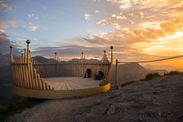 A scenic viewing platform made of wooden panels is situated on a mountain ledge, overlooking a breathtaking landscape of mountain ranges. The sun is setting, casting a warm golden hue across the sky, which contrasts with the darkening mountains in the distance. A person sits on the platform, possibly enjoying the view, with their backpack beside them.