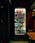 A well-lit refrigerated display filled with various soft drinks and beverages, including bottles and cans. The Coca-Cola branding is prominently displayed at the top of the cooler. The scene is set in a dimly lit room, which contrasts with the bright interior light of the cooler, highlighting the assortment of colorful drinks. Adjacent to the fridge, there is a wooden table and chair.