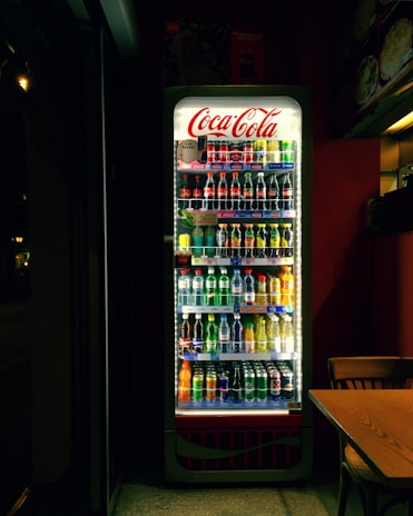 A well-lit refrigerated display filled with various soft drinks and beverages, including bottles and cans. The Coca-Cola branding is prominently displayed at the top of the cooler. The scene is set in a dimly lit room, which contrasts with the bright interior light of the cooler, highlighting the assortment of colorful drinks. Adjacent to the fridge, there is a wooden table and chair.