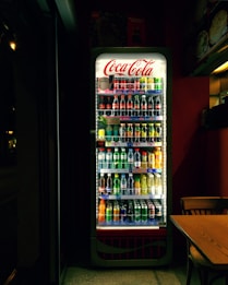 A well-lit refrigerated display filled with various soft drinks and beverages, including bottles and cans. The Coca-Cola branding is prominently displayed at the top of the cooler. The scene is set in a dimly lit room, which contrasts with the bright interior light of the cooler, highlighting the assortment of colorful drinks. Adjacent to the fridge, there is a wooden table and chair.