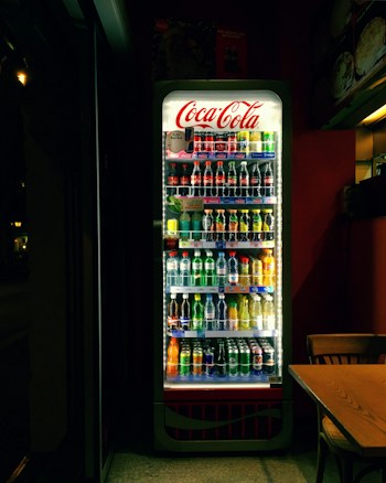 A well-lit refrigerated display filled with various soft drinks and beverages, including bottles and cans. The Coca-Cola branding is prominently displayed at the top of the cooler. The scene is set in a dimly lit room, which contrasts with the bright interior light of the cooler, highlighting the assortment of colorful drinks. Adjacent to the fridge, there is a wooden table and chair.