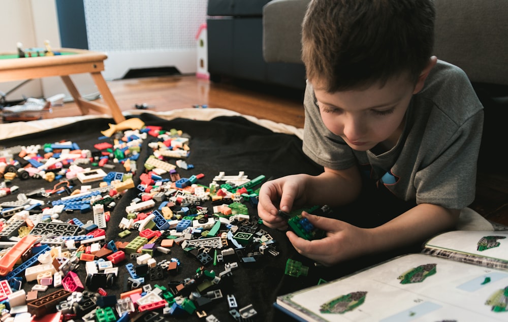 garçon en t-shirt gris à col rond joue aux briques LEGO avec un livre manuel blanc