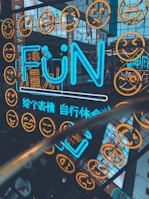 A vibrant and energetic urban scene featuring a neon light display with the word 'FUN' in bright blue, surrounded by numerous yellow smiley face icons on a grid. The colorful signage is reflected on a shiny surface and flanked by additional neon text and advertisements in a bustling cityscape.