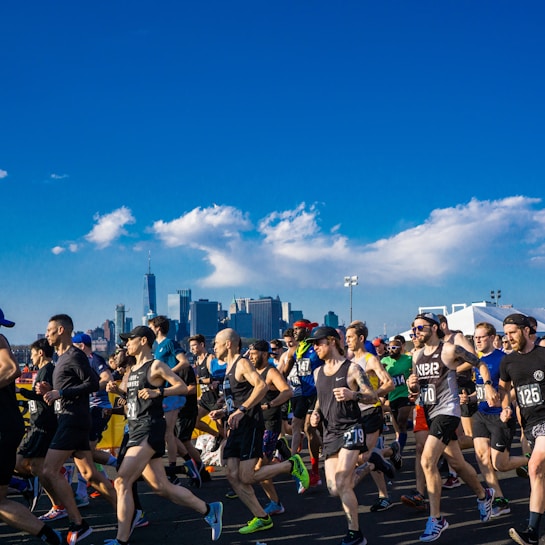 A large group of runners participate in a race, with many wearing numbered bibs. The background features a city skyline with skyscrapers under a clear blue sky with a few white clouds, suggesting an urban setting.
