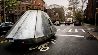 A large metallic, cone-shaped vehicle with a license plate is driving along a city street. Surrounding it are typical urban elements, including other cars, traffic lights, and brick buildings. The atmosphere is overcast and the road surface appears wet.