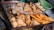 A variety of fried foods on a metal rack, including tofu, shrimp fritters, and vegetable fritters. Tongs rest on top of the food, and there are bowls and a pot in the background.