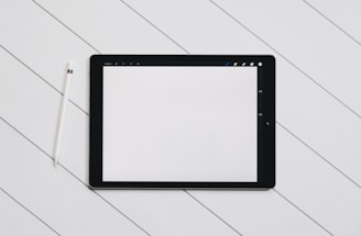 A digital tablet with a blank white screen sits on a light wooden surface. To the left of the tablet, there is a white stylus placed parallel to it. The setting is simple and minimalistic, emphasizing the technology and design.