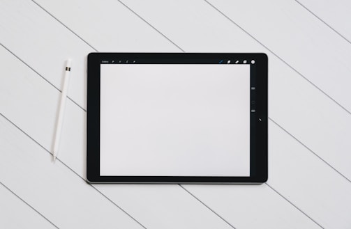 A digital tablet with a blank white screen sits on a light wooden surface. To the left of the tablet, there is a white stylus placed parallel to it. The setting is simple and minimalistic, emphasizing the technology and design.