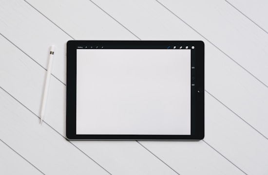 A digital tablet with a blank white screen sits on a light wooden surface. To the left of the tablet, there is a white stylus placed parallel to it. The setting is simple and minimalistic, emphasizing the technology and design.