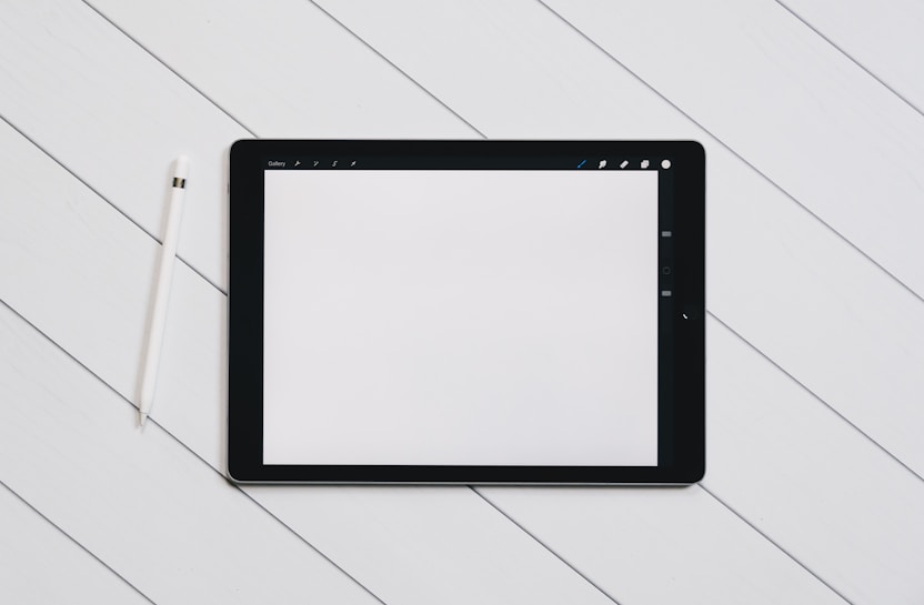 A digital tablet with a blank white screen sits on a light wooden surface. To the left of the tablet, there is a white stylus placed parallel to it. The setting is simple and minimalistic, emphasizing the technology and design.