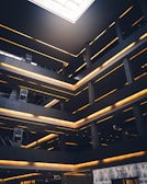 A modern architectural interior featuring multiple levels with glowing linear lights. The space has a geometric design with dark grey and black tones, contrasted by warm yellow lighting. There are signs with the word 'SOHO' and a high-end storefront labeled 'Dior' on the ground level. A large skylight at the top allows natural light to illuminate part of the space.