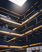 A modern architectural interior featuring multiple levels with glowing linear lights. The space has a geometric design with dark grey and black tones, contrasted by warm yellow lighting. There are signs with the word 'SOHO' and a high-end storefront labeled 'Dior' on the ground level. A large skylight at the top allows natural light to illuminate part of the space.
