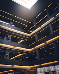 A modern architectural interior featuring multiple levels with glowing linear lights. The space has a geometric design with dark grey and black tones, contrasted by warm yellow lighting. There are signs with the word 'SOHO' and a high-end storefront labeled 'Dior' on the ground level. A large skylight at the top allows natural light to illuminate part of the space.