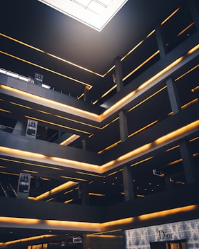 A modern architectural interior featuring multiple levels with glowing linear lights. The space has a geometric design with dark grey and black tones, contrasted by warm yellow lighting. There are signs with the word 'SOHO' and a high-end storefront labeled 'Dior' on the ground level. A large skylight at the top allows natural light to illuminate part of the space.
