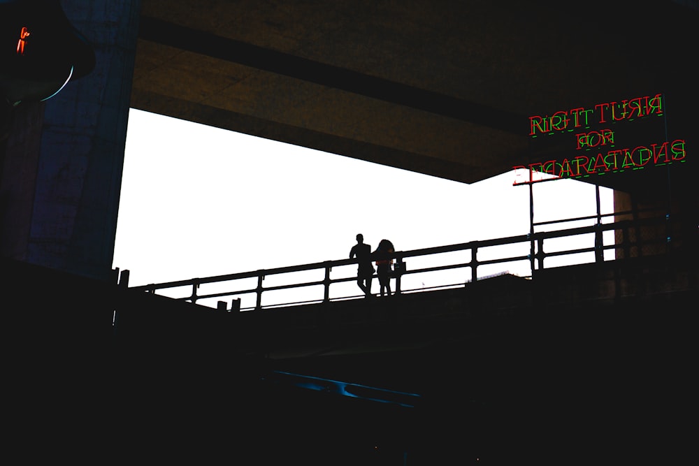 silhouette photo of two person standing beside metal railings near LED signage