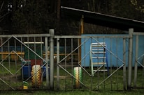 A playground scene with metal fencing featuring diamond-shaped patterns in the foreground. Behind the fence is a structure resembling a playhouse or shed painted in light blue. A colorful playground cart with yellow tires and multicolored benches can also be seen. The ground is covered with grass and there are trees in the background.