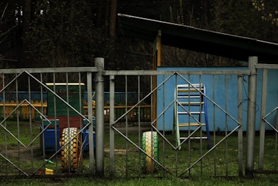 A playground scene with metal fencing featuring diamond-shaped patterns in the foreground. Behind the fence is a structure resembling a playhouse or shed painted in light blue. A colorful playground cart with yellow tires and multicolored benches can also be seen. The ground is covered with grass and there are trees in the background.