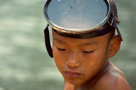 A young boy looks downwards, wearing a head strap with a circular, clear plastic lens against a blurred background. His expression appears contemplative.