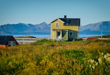An old yellow house with a black roof is situated in a grassy field with mountains and the sea in the background. Another small red building is visible to the left, surrounded by a serene and natural landscape.