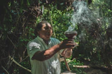 A wise shaman gently holding palo santo, with forest greenery blurring softly in the background.