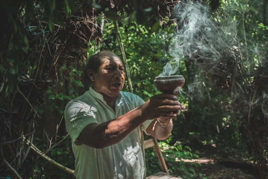 A person in a white shirt stands in a lush forest, holding a smoking vessel. Light filters through the dense greenery, highlighting the smoke that rises from the vessel. The individual appears focused, suggesting an act of ritual or meditation.