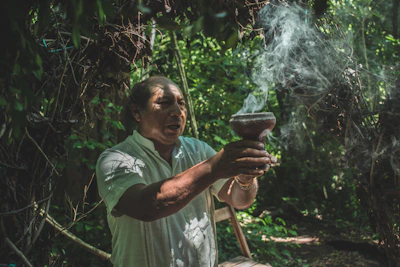 A wise shaman gently holding palo santo, with forest greenery blurring softly in the background.