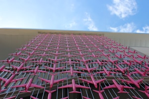 An artistic installation with numerous small pink airplane models arranged in a grid pattern against an exterior building wall. The sky with clouds is visible above, contrasting the vivid planes against the light-colored structure.