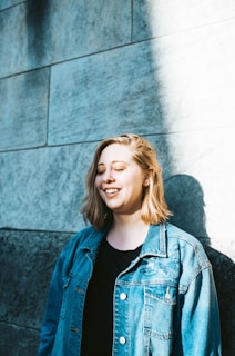 A person with short blonde hair smiles while standing against a textured stone wall. Sunlight casts a bright shadow on the wall, highlighting the person's face and denim jacket.