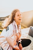 A person is smiling joyfully in a bright, sunny outdoor setting, wearing a light-colored jacket with a letterman style design. The background features soft, blurred hills and clear skies, enhancing the warm and cheerful ambiance.