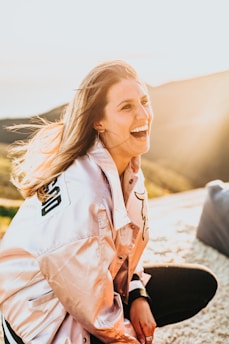 A person is smiling joyfully in a bright, sunny outdoor setting, wearing a light-colored jacket with a letterman style design. The background features soft, blurred hills and clear skies, enhancing the warm and cheerful ambiance.