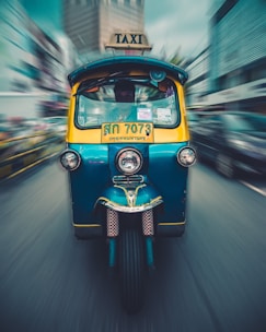 A colorful tuk-tuk taxi is moving at speed on a street, with a blurred urban background conveying motion. The vehicle is blue and yellow, with a bright TAXI sign on top and a license plate in the local script. Buildings and other vehicles in the background are blurred, suggesting a bustling city environment.