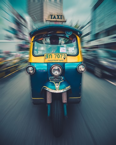 A colorful tuk-tuk taxi is moving at speed on a street, with a blurred urban background conveying motion. The vehicle is blue and yellow, with a bright TAXI sign on top and a license plate in the local script. Buildings and other vehicles in the background are blurred, suggesting a bustling city environment.