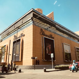 A modern building with a tan exterior and large display advertisements on the walls. The name 'El Palacio de Hierro' is prominently featured. Multiple signs and a streetlight are visible in front. Several people and a motorcycle are present near the entrance.