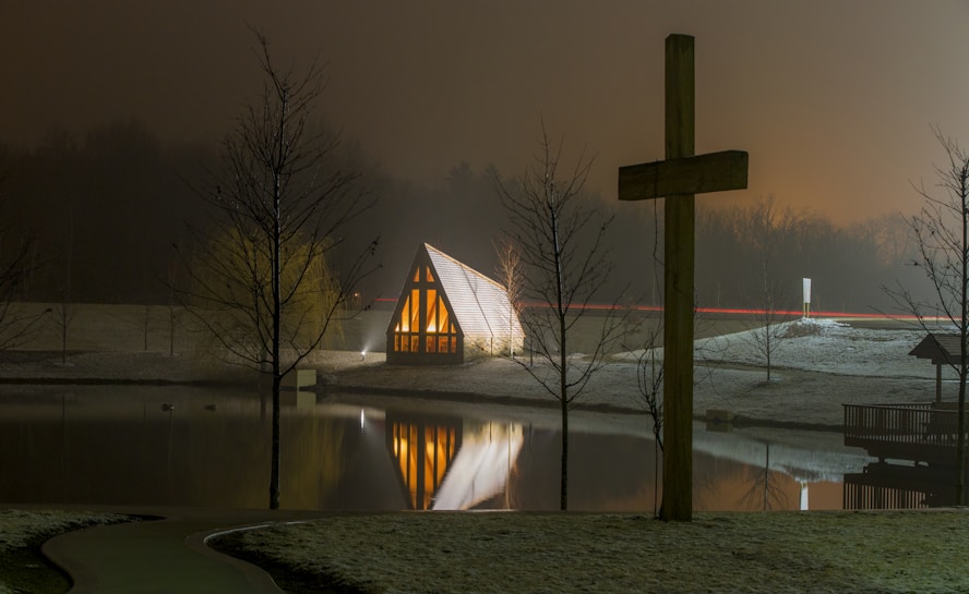 A small chapel-like structure with illuminated windows is reflected in a calm body of water. A large wooden cross stands prominently on the right side, and the area is surrounded by leafless trees. The sky has an overcast, misty appearance, contributing to a serene and contemplative atmosphere.