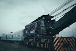 An old, weathered railway crane is connected to cargo train cars on a cloudy day. The crane's dark teal and rusty exterior suggests long-term use and exposure to the elements. Two individuals are seen near the cabin of the crane, with one standing on the steps, giving a sense of exploration or maintenance work. The barren landscape and overcast sky contribute to a moody and somber atmosphere.
