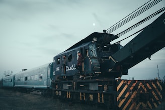 An old, weathered railway crane is connected to cargo train cars on a cloudy day. The crane's dark teal and rusty exterior suggests long-term use and exposure to the elements. Two individuals are seen near the cabin of the crane, with one standing on the steps, giving a sense of exploration or maintenance work. The barren landscape and overcast sky contribute to a moody and somber atmosphere.