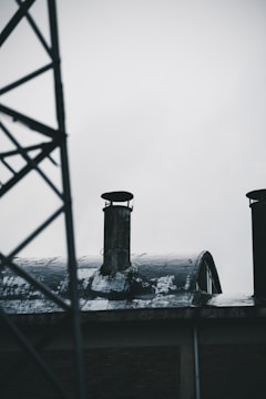 An industrial setting with two weathered chimneys on a corrugated metal roof, partly covered with snow. A structural metal framework is visible on the left side of the image, against an overcast sky.