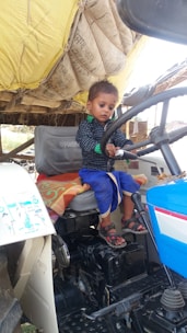 A young child is sitting on the driver's seat of a tractor, holding the steering wheel. The tractor has a blue and white color scheme, and there is a large bundle of hay or similar material tied at the back. The child is wearing a black and blue outfit with sandals.