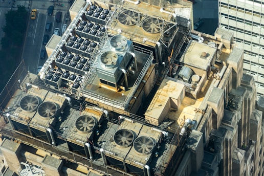 A detailed aerial view of an industrial rooftop displaying numerous ventilation systems and fans. The setup includes multiple large fans enclosed by metal railings, arranged neatly along the roof’s surface. The surrounding architecture features geometric shapes and utilitarian design elements, indicative of a commercial or industrial building. The streets and structures below indicate an urban environment, with vehicles and tall buildings partially visible.