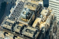 A detailed aerial view of an industrial rooftop displaying numerous ventilation systems and fans. The setup includes multiple large fans enclosed by metal railings, arranged neatly along the roof&rsquo;s surface. The surrounding architecture features geometric shapes and utilitarian design elements, indicative of a commercial or industrial building. The streets and structures below indicate an urban environment, with vehicles and tall buildings partially visible.