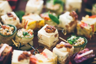 A variety of elegant appetizers are displayed, including small cubes of cheese topped with walnuts, rolls filled with creamy mixtures, and cups containing grain salads and greens. The presentation is artistic and colorful, with each appetizer meticulously arranged and garnished.