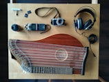 An overhead view of a wooden table with various items laid out including a stringed musical instrument, a digital audio recorder, a camera, headphones, a lens, and memory cards. The musical instrument appears to be a zither, featuring numerous strings and a wooden body. The digital audio recorder and camera are compact, with a lens cap nearby. Large over-ear headphones are also present with their cable coiled neatly.