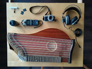 An overhead view of a wooden table with various items laid out including a stringed musical instrument, a digital audio recorder, a camera, headphones, a lens, and memory cards. The musical instrument appears to be a zither, featuring numerous strings and a wooden body. The digital audio recorder and camera are compact, with a lens cap nearby. Large over-ear headphones are also present with their cable coiled neatly.