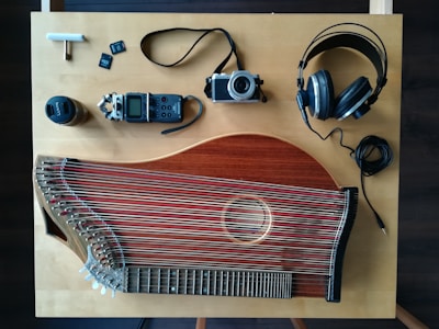 An overhead view of a wooden table with various items laid out including a stringed musical instrument, a digital audio recorder, a camera, headphones, a lens, and memory cards. The musical instrument appears to be a zither, featuring numerous strings and a wooden body. The digital audio recorder and camera are compact, with a lens cap nearby. Large over-ear headphones are also present with their cable coiled neatly.
