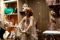 A fashion window display features an elegantly dressed mannequin seated amidst a luxurious setup. The mannequin wears a detailed lace dress and jewelry, surrounded by a variety of high-end handbags, accessories, and decorative elements. The setting conveys opulence with a vintage-styled chair, ornate shelving, and a warm color palette.