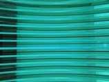 Several horizontal turquoise glass panes are stacked in parallel, creating a layered and reflective surface. The light passing through the glass gives it a luminous and modern appearance.