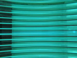 Several horizontal turquoise glass panes are stacked in parallel, creating a layered and reflective surface. The light passing through the glass gives it a luminous and modern appearance.