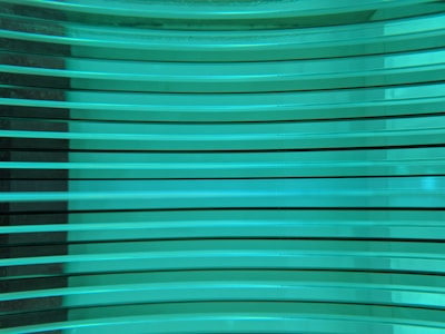 Several horizontal turquoise glass panes are stacked in parallel, creating a layered and reflective surface. The light passing through the glass gives it a luminous and modern appearance.